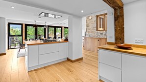 A modern kitchen with a central island, wooden flooring, and large windows offering a view of greenery. - Mercia House - sleeps 8 guests  in 4 bedrooms (Cheltenham)