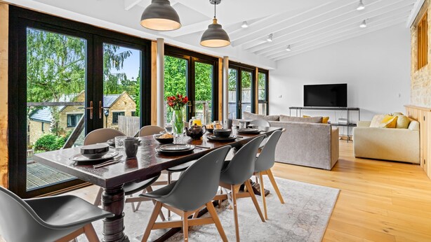 A modern dining area with a large table set for breakfast, surrounded by large windows offering a view of the outdoors. - Mercia House - sleeps 8 guests  in 4 bedrooms (Cheltenham)