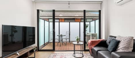 A modern living room with a flat-screen TV, a leather sofa, a patterned rug, and a sliding glass door leading to an outdoor patio area.