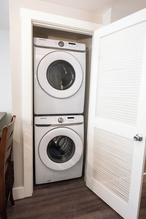 Una lavandería con dos lavadoras apiladas, una silla de madera y una puerta blanca con rejilla de ventilación.