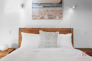 Ein Schlafzimmer mit einem hölzernen Kopfteil, weißer Bettwäsche, einem gemusterten Kopfkissen und Strandbilder an der Wand.