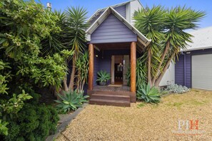 A house with a wooden porch, surrounded by tropical plants and a gravel driveway. - Beachwood Retreat for Ultimate Relaxation (Cowes)