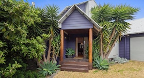 A house with a wooden porch, surrounded by tropical plants and a gravel driveway.