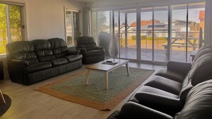Ein geräumiges Wohnzimmer mit einer großen Couch, einem kleineren Sessel und einem Couchtisch. Ein Deckenventilator und große Fenster bieten einen Blick ins Freie.