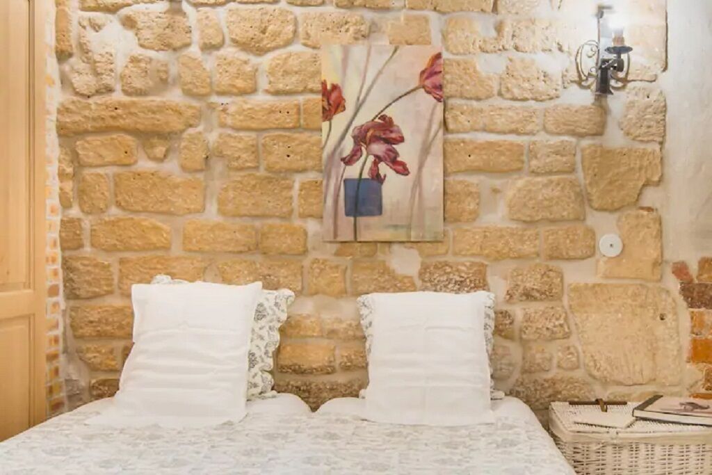 Une pièce avec un mur en pierre, un lit recouvert de draps blancs et un vase décoratif rempli de fleurs.