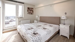 A bedroom with a large bed, a nightstand, a dresser, a window with a view of buildings, and a radiator. - Beautiful apartment in Oostende (Oostende)