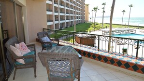 Een balkon met rieten stoelen en een glazen tafel, met uitzicht op een zwembad en een hotelgebouw.