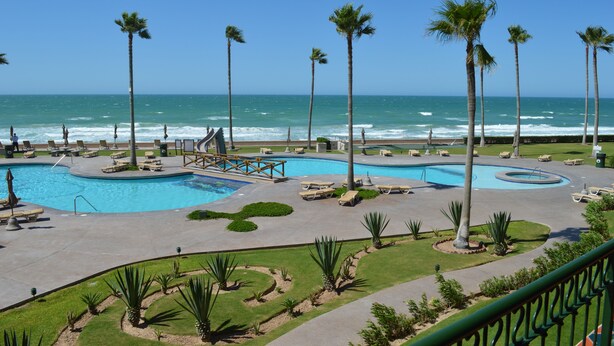 Zwembad van een resort met palmbomen, een houten brug en uitzicht op de oceaan.