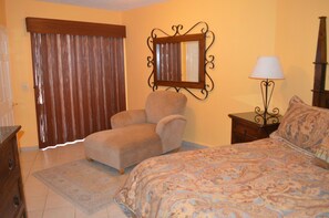 Quarto com cama, cadeira, mesa de cabeceira, luminária, espelho e janela com cortinas.