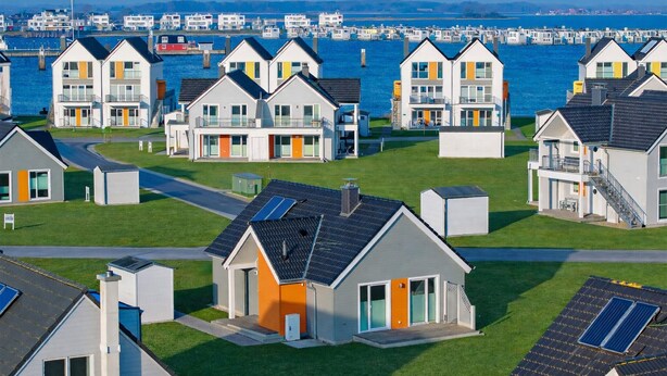 Zona residencial costera con casas modernas que tienen balcones, paneles solares y un puerto deportivo al fondo.