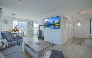 A modern living room with a large flat-screen TV, a dining area with a table and chairs, and a kitchen area with a cart and a hanging net.