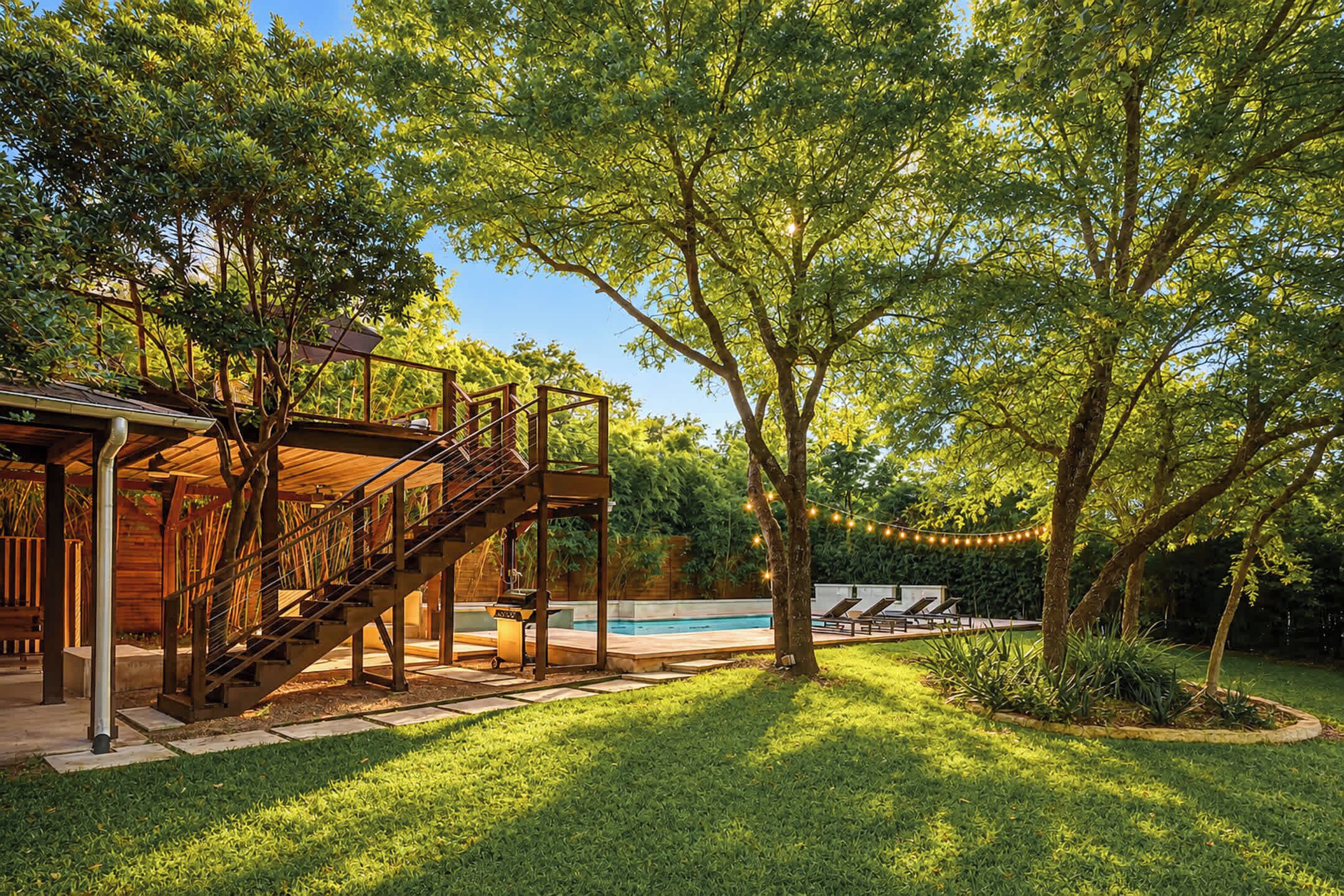 A wooden deck with a staircase leading to a covered area, a swimming pool, and a well-maintained lawn.