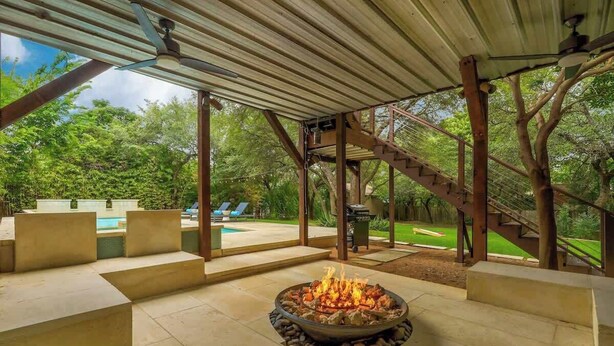 Un patio exterior con una fogata, asientos y vistas a una piscina y jardín.