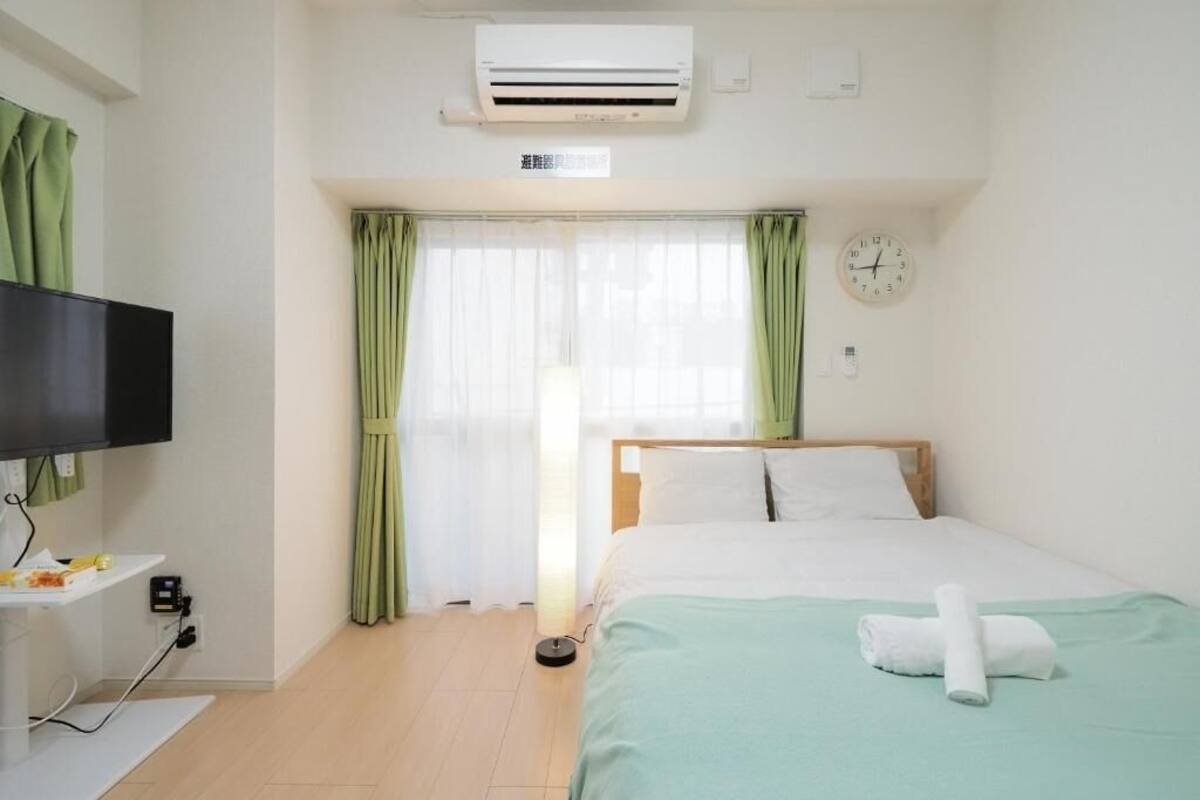 A hotel room with a bed, a TV, an air conditioner, a clock, and a window with curtains.