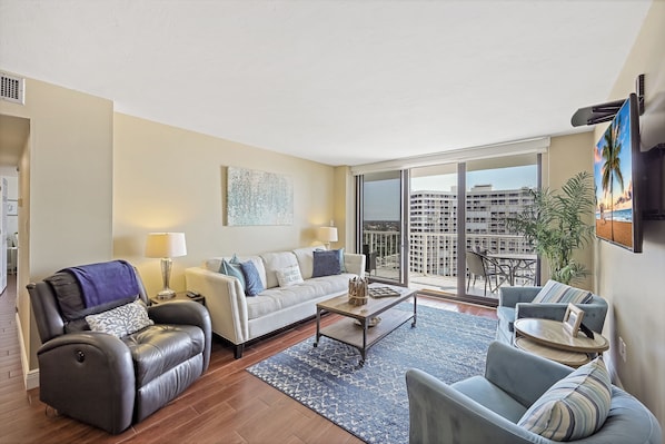 A modern living room with a sofa, armchair, and coffee table, a large window with a view of buildings, and a balcony with plants.