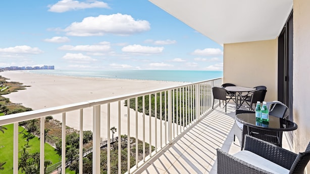 A balcony with a view of a beach, a table with chairs, and two bottles of water. - Relaxing Beachfront Condo | Pool & Ocean Views (Marco Island)