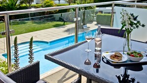 A balcony with a glass table, two wine glasses, a bottle, a bowl of salad, and a plate with bread.