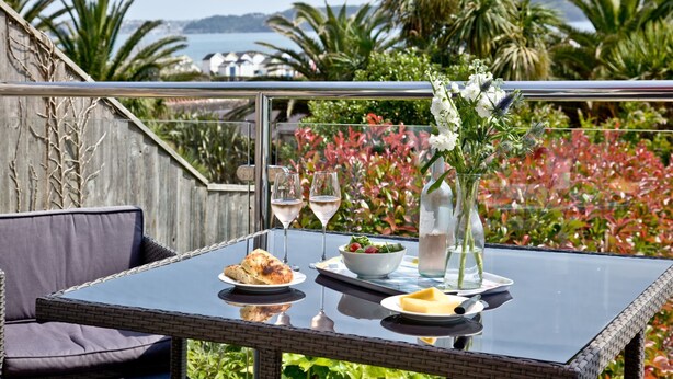A table set with a glass of white wine, a bowl of salad, a plate of cheese, and a bottle of water, with a view of the sea and palm trees in the background.