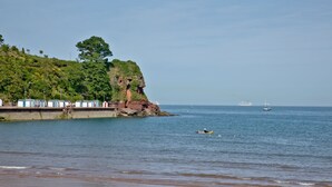 A beach with a rocky cliff, a pier with beach huts, and a yacht in the distance.
