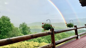 Eine Holzterrasse mit Geländer, die auf eine üppige grüne Landschaft und einen Regenbogen am Himmel blickt.