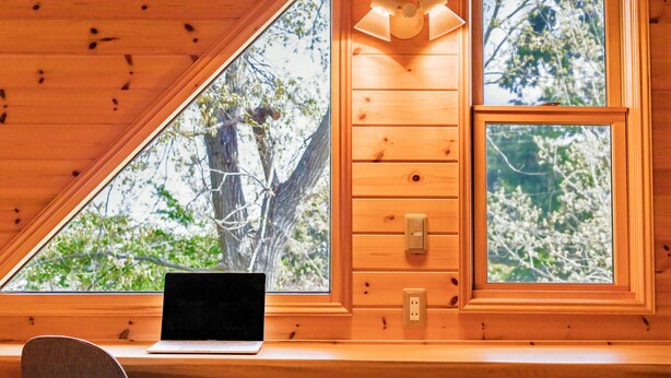 A wooden cabin interior with a laptop on a desk, large windows with a view of trees, and a wall-mounted light fixture. - Izu House Luxury Log with Cozy Fireplace (Ito)