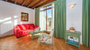 Una sala de estar con un sofá rojo, una mesa de centro de cristal, un sillón con estampado floral, una mesita auxiliar verde con un cuenco decorativo y un ventanal con cortinas verdes.