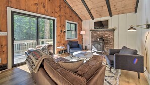 A cozy living room with a brown leather sofa, a gray armchair, and a coffee table. There is a fireplace with a brick surround and a large window overlooking a deck. - Sleek Cabin w/ Deck, 8 Miles to Mount Snow & Hikes (Wilmington)