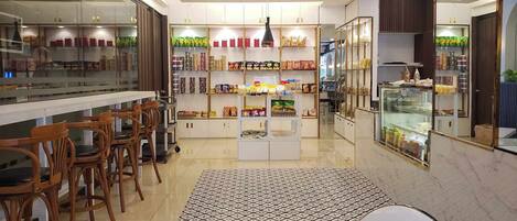 A modern cafe interior with wooden chairs, a patterned rug, and shelves stocked with snacks and beverages.