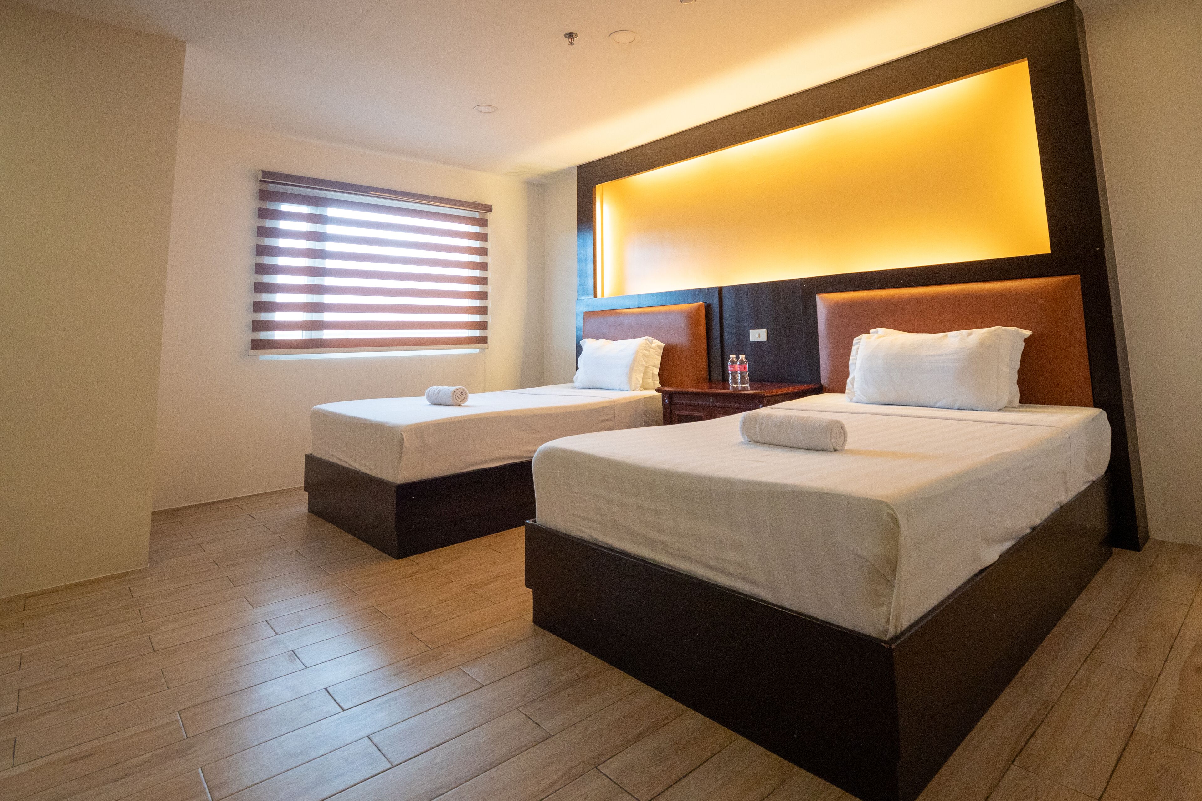 A hotel room with two beds, wooden flooring, a large illuminated wall panel, and a window with blinds.