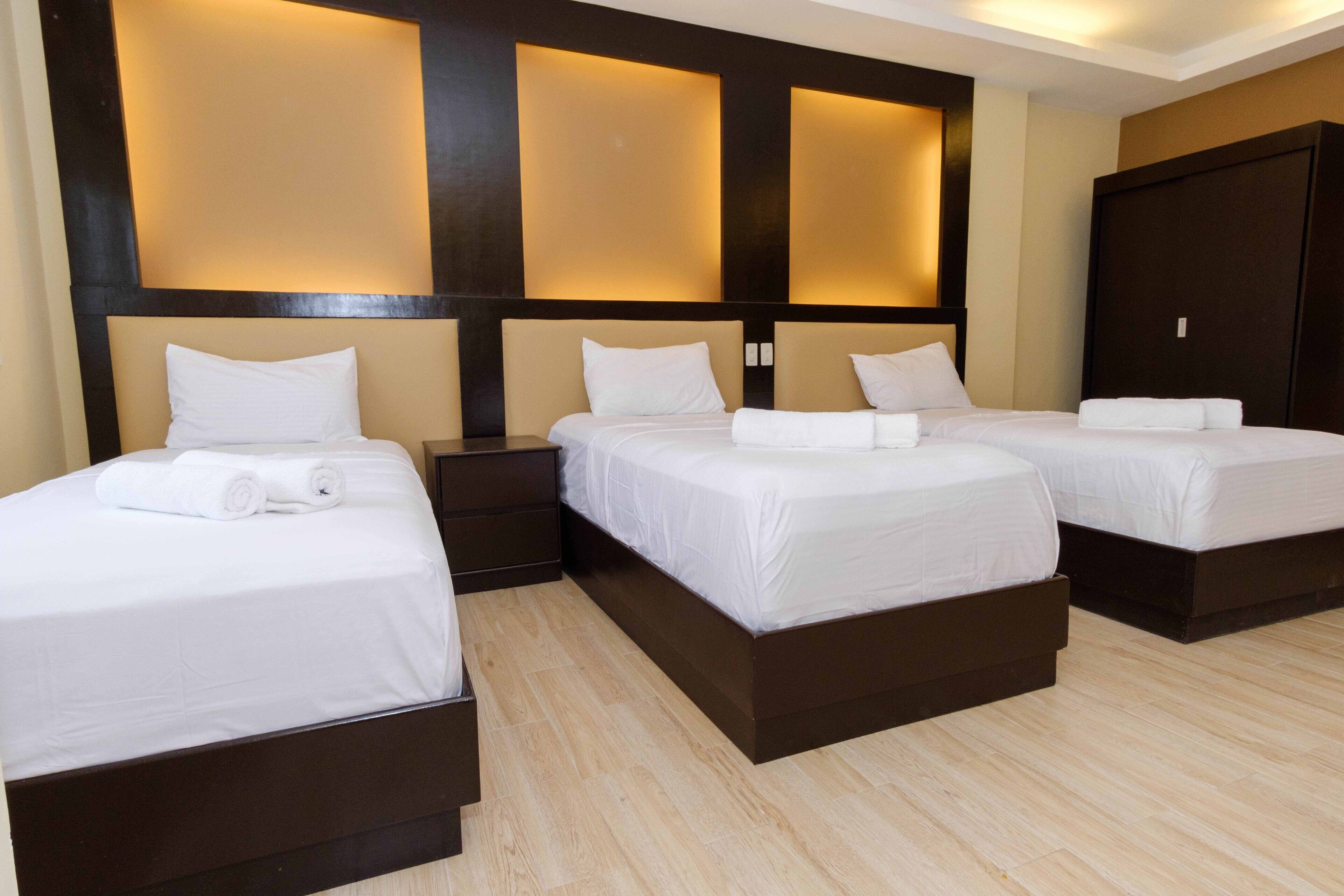 A hotel room with three beds, each with white linens and a dark headboard. The room has a modern design with a wooden floor and a large wardrobe.