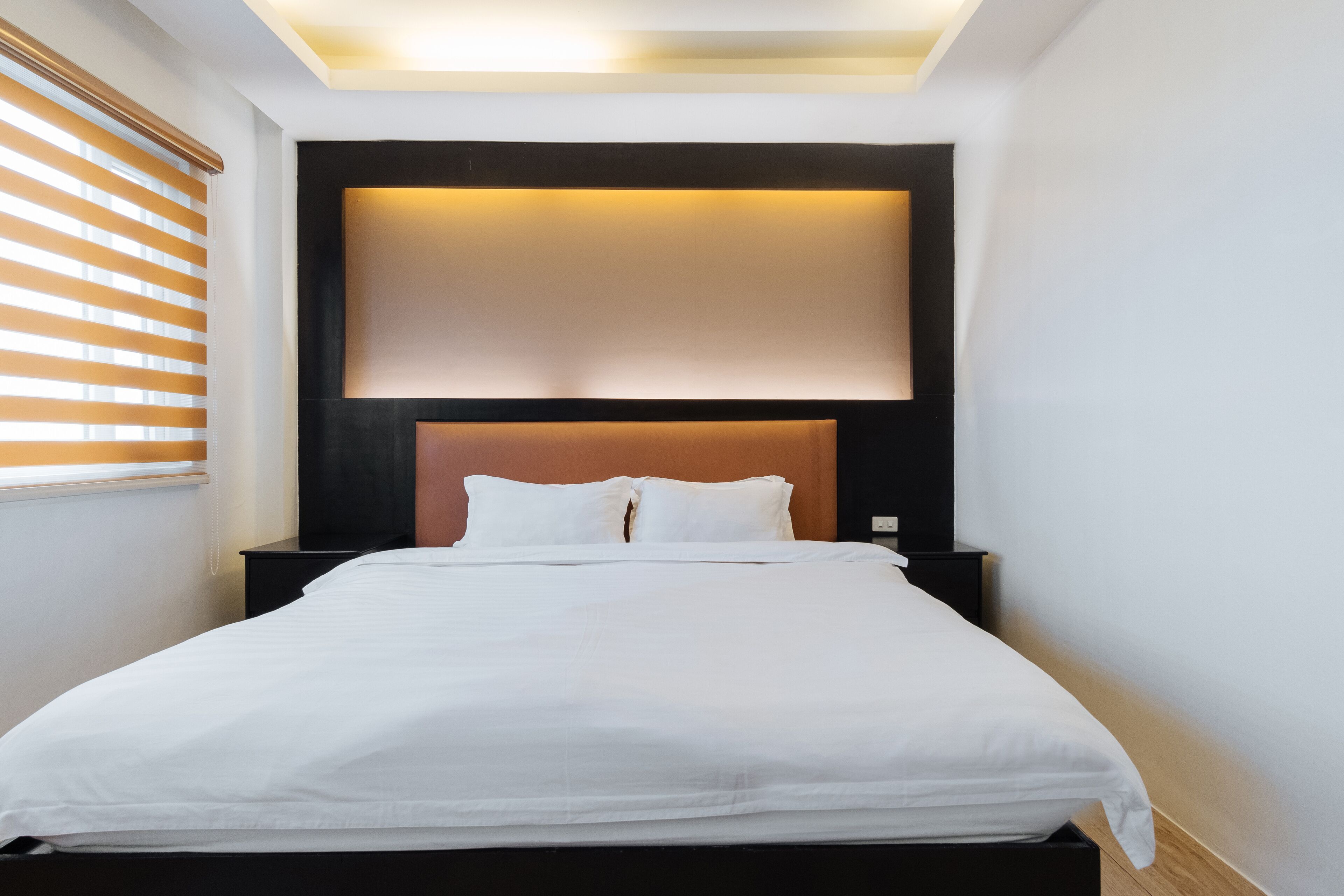 A modern hotel room with a large bed, a headboard with built-in lighting, a bedside table, and a window with blinds.