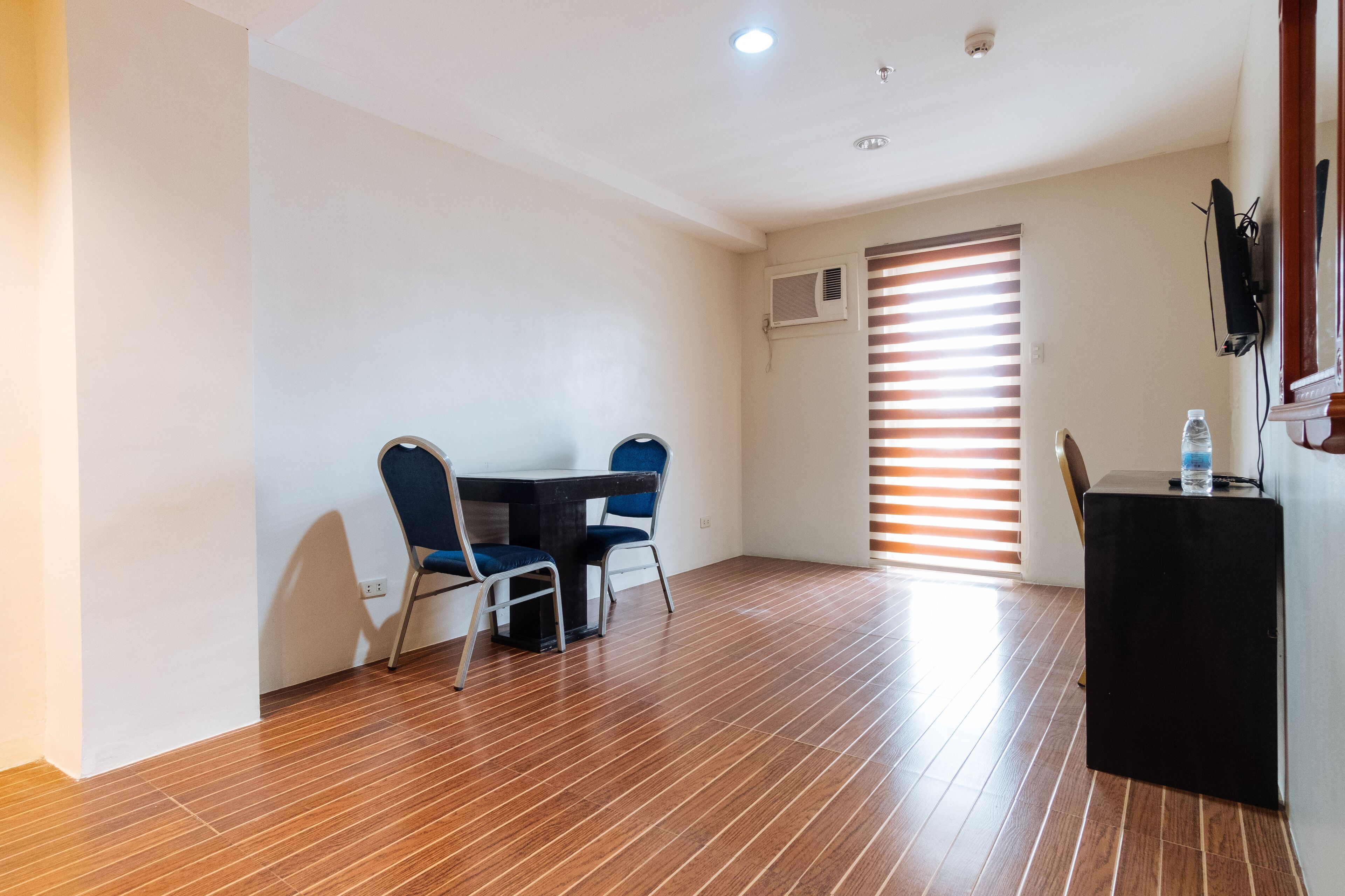 A room with a wooden floor, a table with two chairs, a black side table, and a window with blinds.