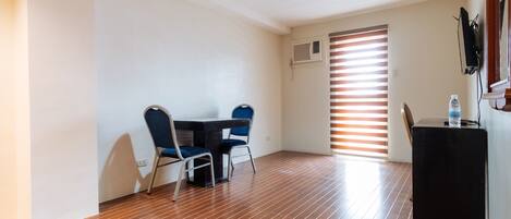 A room with a wooden floor, a table with two chairs, a black side table, and a window with blinds.