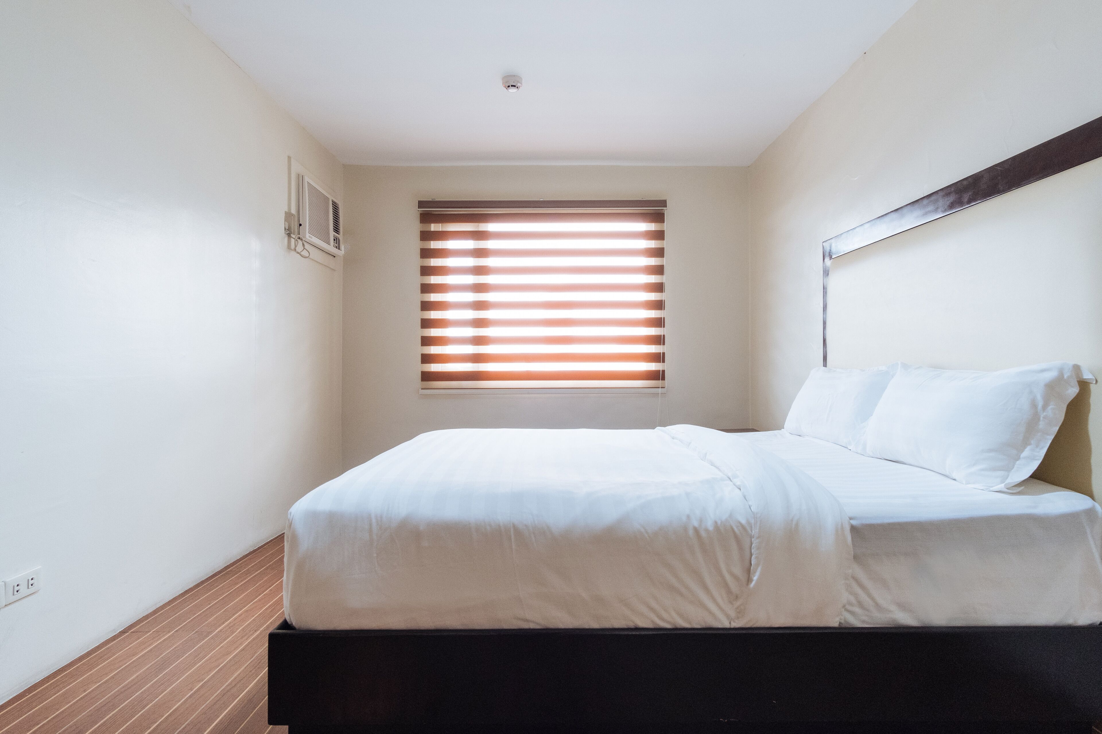 A bedroom with a bed, a window with blinds, a ceiling light, and a wall-mounted air conditioning unit.