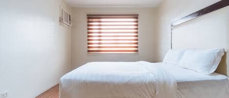 A bedroom with a bed, a window with blinds, a ceiling light, and a wall-mounted air conditioning unit.