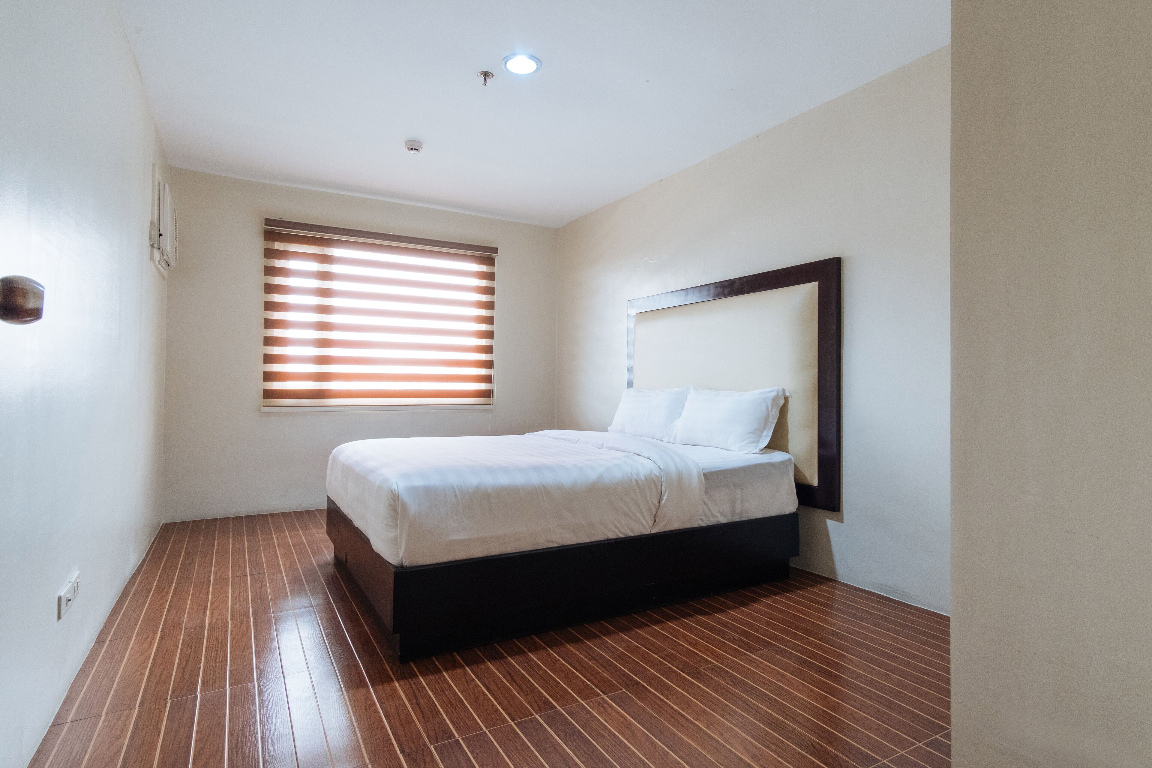 Une chambre à coucher avec un lit, un plancher en bois, une fenêtre avec des stores et un luminaire au plafond.