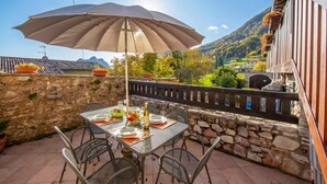 Un espace de restauration en plein air avec une table dressée pour deux personnes, un grand parasol, des murs en pierre et une vue sur une montagne.