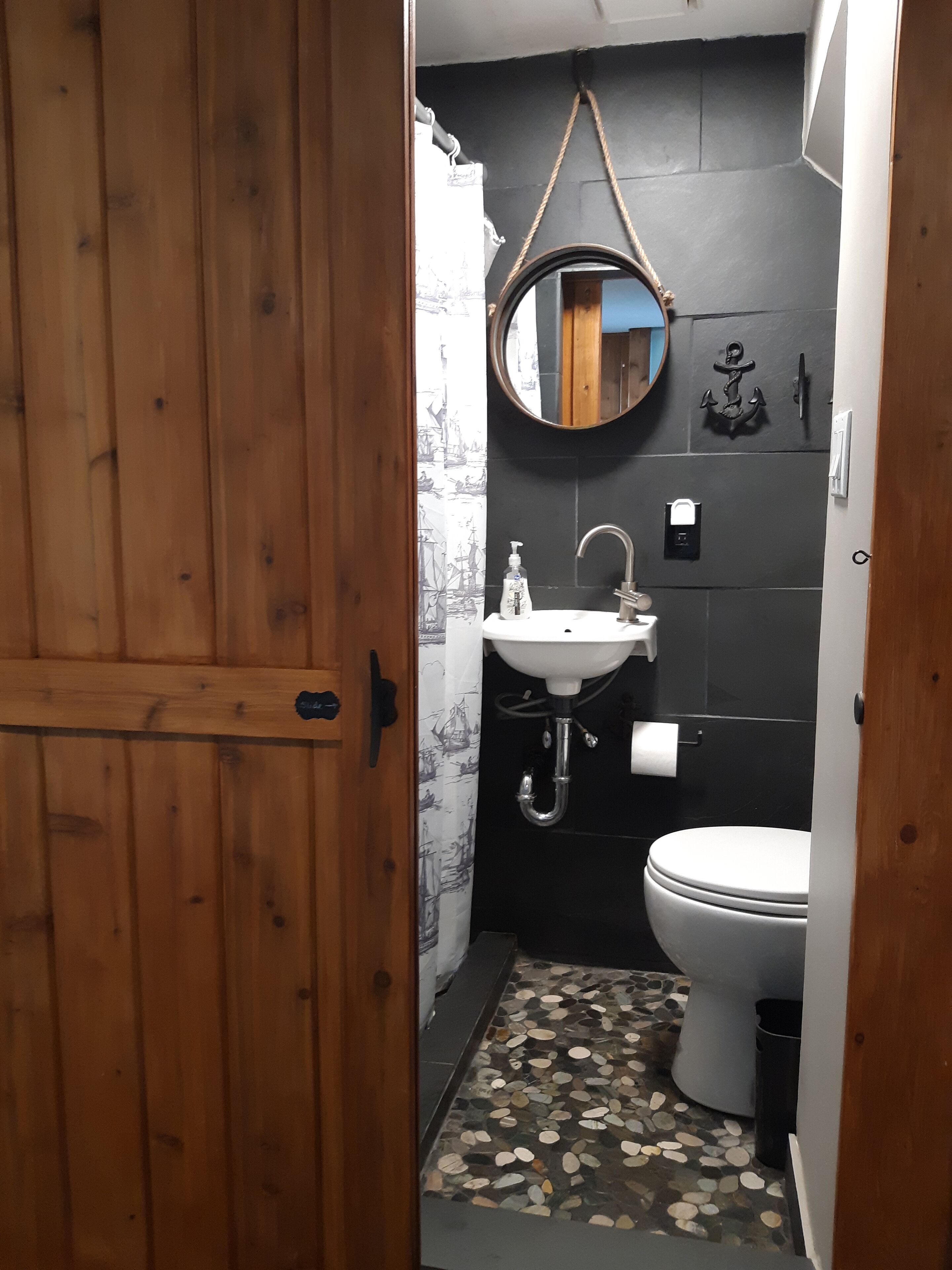 A compact bathroom with a wooden door, a round mirror, a white sink, a toilet, and a patterned floor.