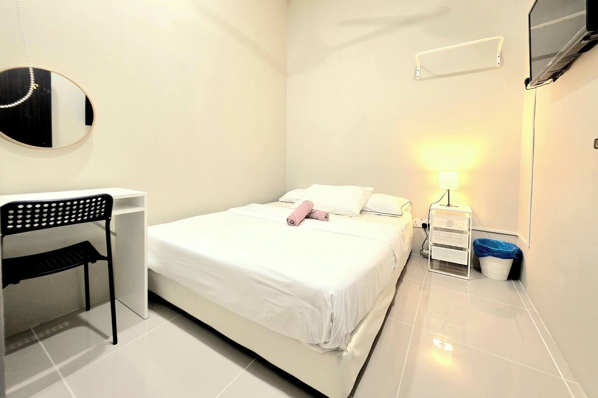 A clean, modern hotel room with a bed, a desk, a chair, a TV, a lamp, and a trash bin.