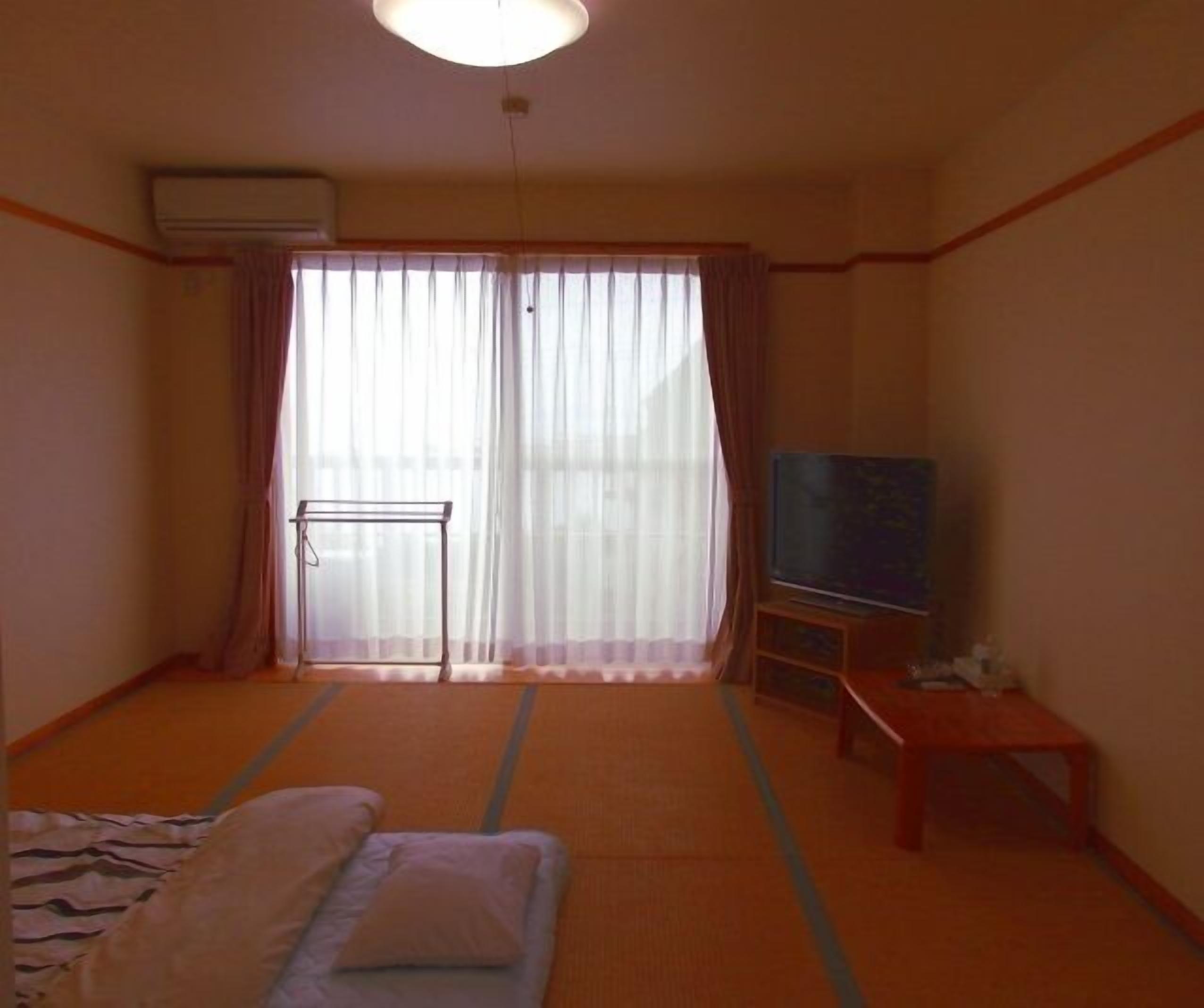 A room with tatami mats, a bed with pillows, a TV on a wooden stand, and a sliding door with curtains.