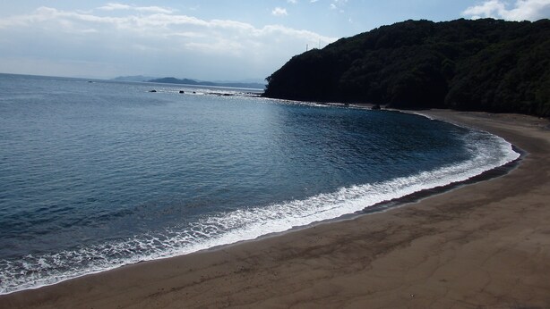 Una playa con una orilla curva, aguas tranquilas y una colina boscosa al fondo.