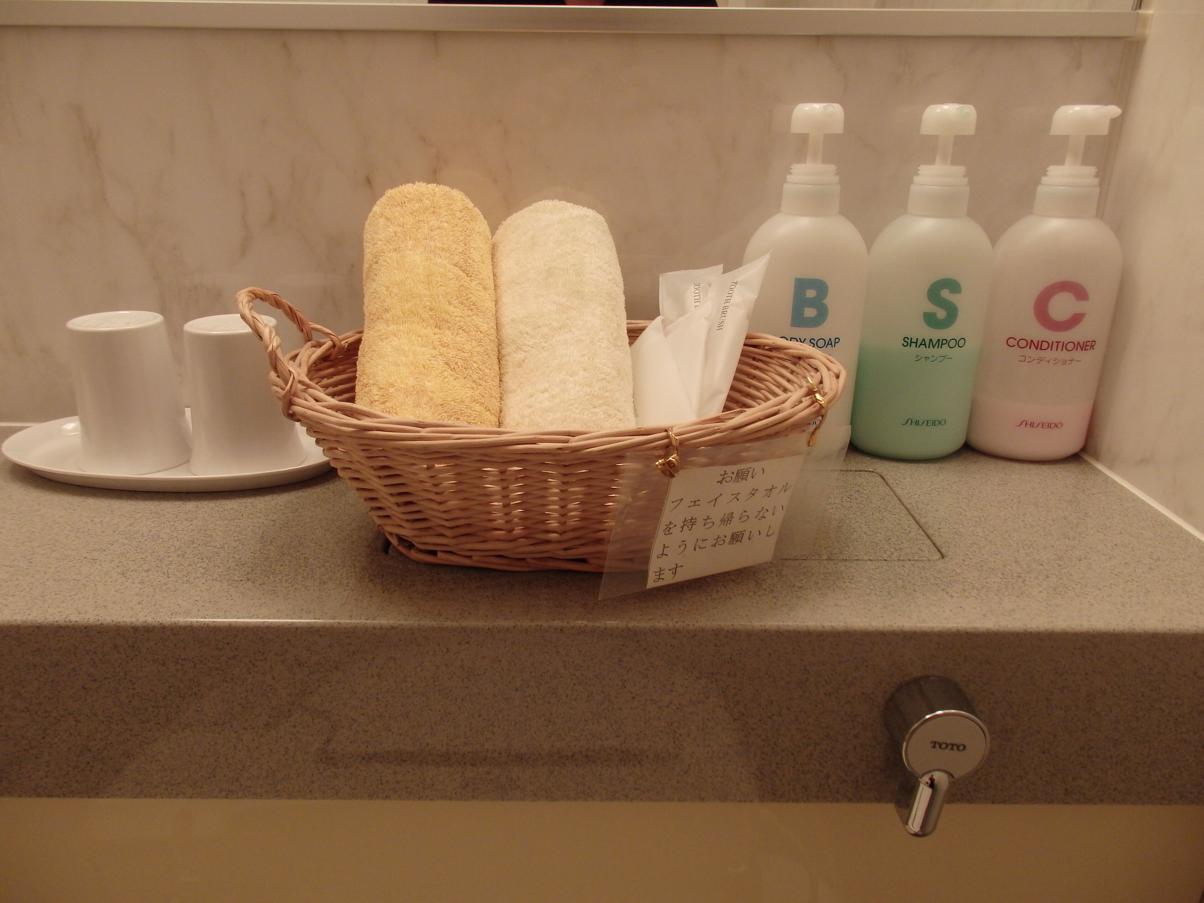 A bathroom counter with a wicker basket containing towels, a soap dispenser, and a small white plate with two cups.