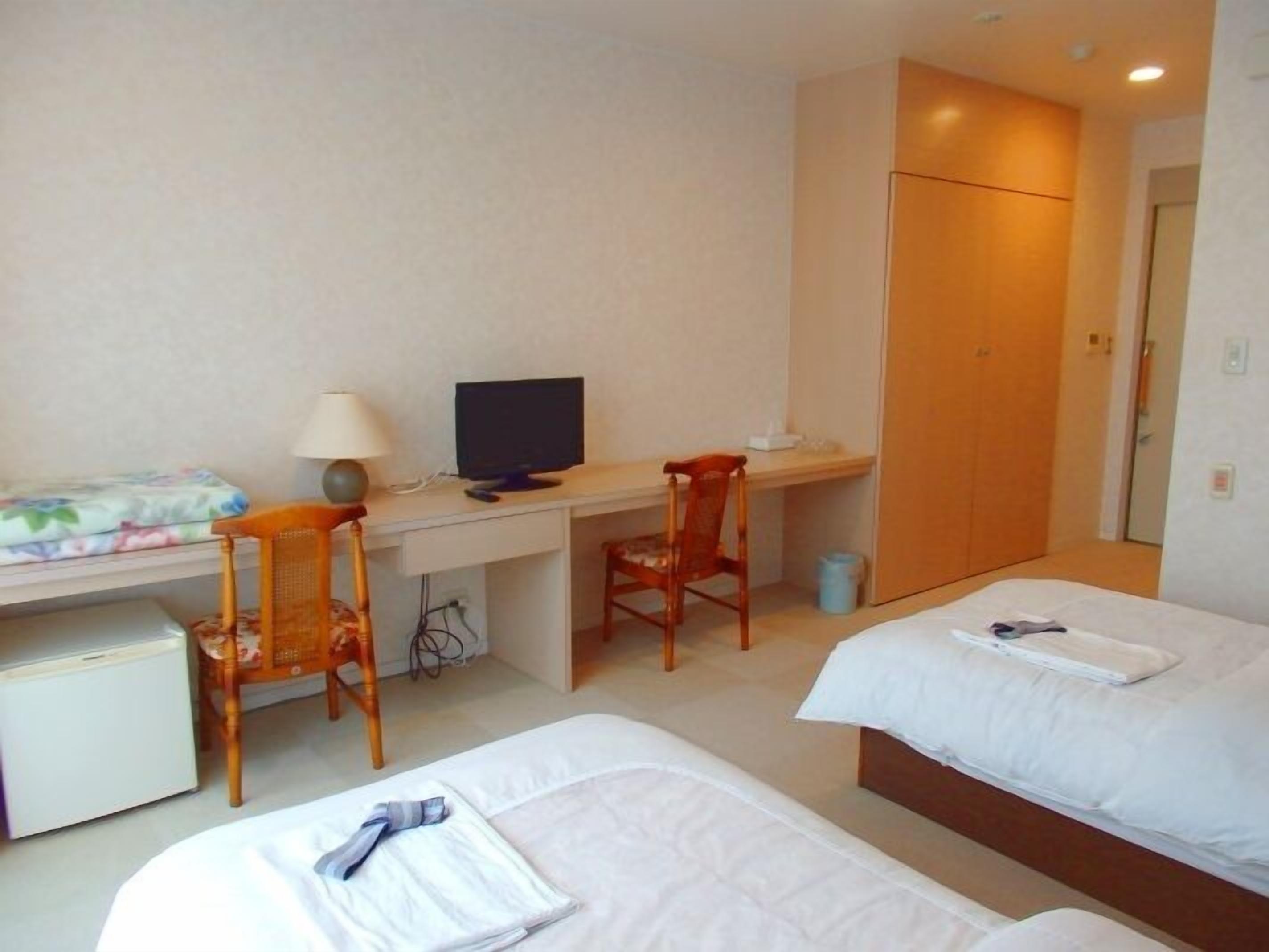 A hotel room with a desk, two beds, a chair, a small refrigerator, and a door.