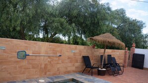 A swimming pool with steps leading into it, surrounded by a brick wall. There are two black lounge chairs and a thatched umbrella.