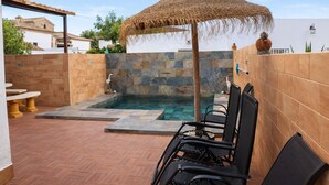 A swimming pool area with a stone wall, a thatched umbrella, and black lounge chairs.