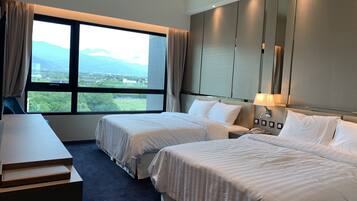 A hotel room with two beds, a desk, a lamp, and a large window with a view of greenery and buildings.