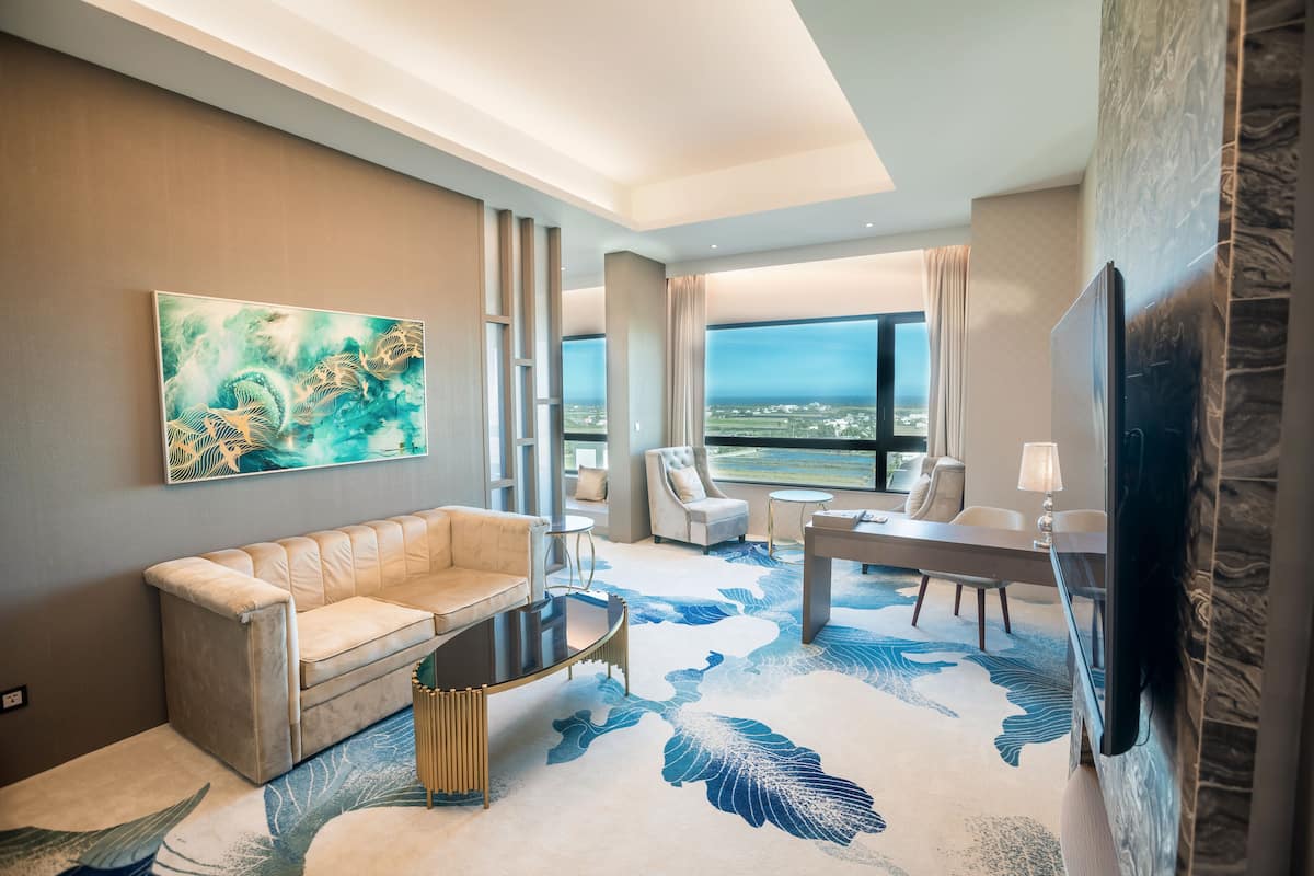 Ein modernes Hotelzimmer mit großem Fenster, einem beigefarbenen Sofa, einem Couchtisch, einem Esstisch mit Stühlen und einem Flachbildfernseher.