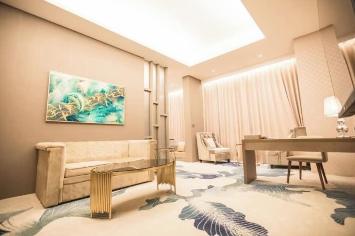 A modern hotel room with a beige sofa, a glass coffee table, a dining table with chairs, and a large abstract painting on the wall.