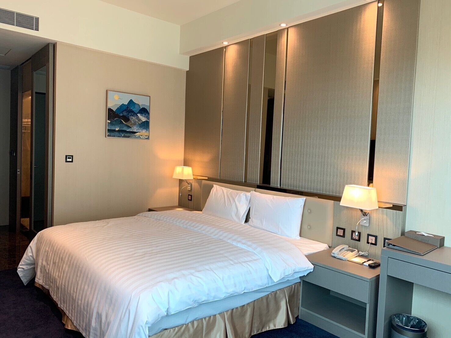 A hotel room with a large bed, two bedside lamps, a telephone, and a small desk with a trash bin.