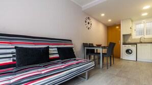 A modern living space with a striped sofa, a dining table with chairs, a kitchen area, and a wall clock. - Apartment Alda by Interhome (Segur de Calafell)